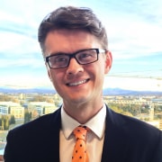 A professional headshot of a smiling man with glasses, wearing a suit and an orange patterned tie, standing in front of a window with a view of a cityscape.