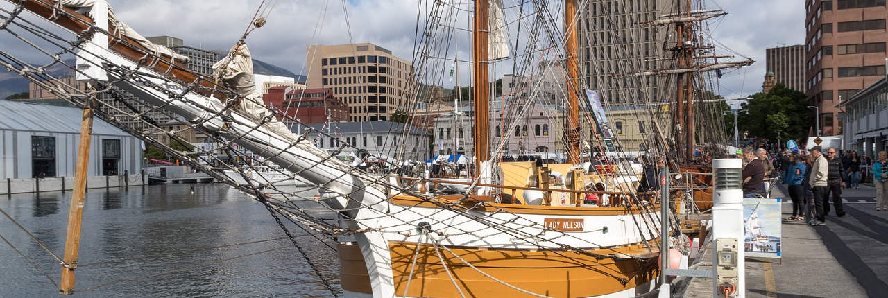 A panoramic view of a harbor with a vintage sailing ship docked at the quayside. The ship's bow is in the foreground, with intricate rigging and a wooden hull, named "LADY NELSON." People are visible along the dock, indicating a lively waterfront scene, with modern buildings in the background under a cloudy sky.