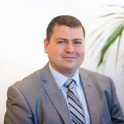 A professional headshot of a man in a grey suit and blue tie, likely for a corporate profile.