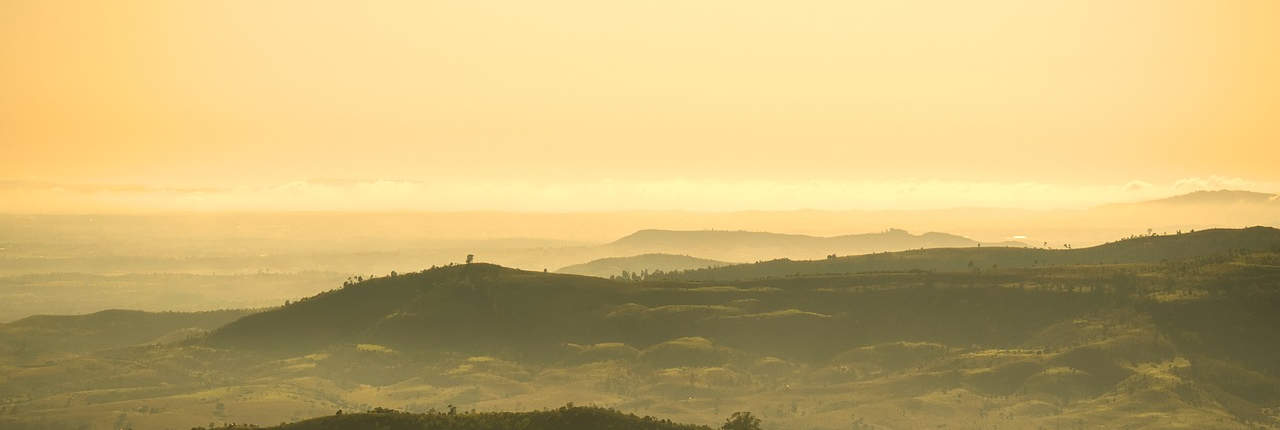 A panoramic view of a landscape bathed in the golden light of sunrise or sunset, featuring rolling hills with a figure standing atop one hill, highlighting the beauty and tranquility of the scene.