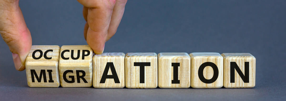 A close-up of a hand completing a row of wooden blocks with letters on them that spell out "IMMIGRATION," with the hand placing the last letter. The focus is on the concept of completing or building up immigration, likely in the context of policy or discussion.