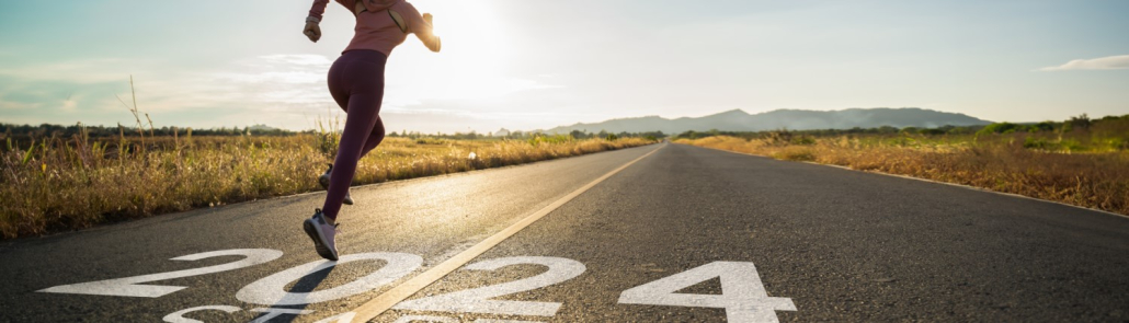 A person sprinting on a road with "2024" painted on the surface, indicating the year, with an early morning or late afternoon rural landscape in the background, featuring a clear sky and distant mountains.