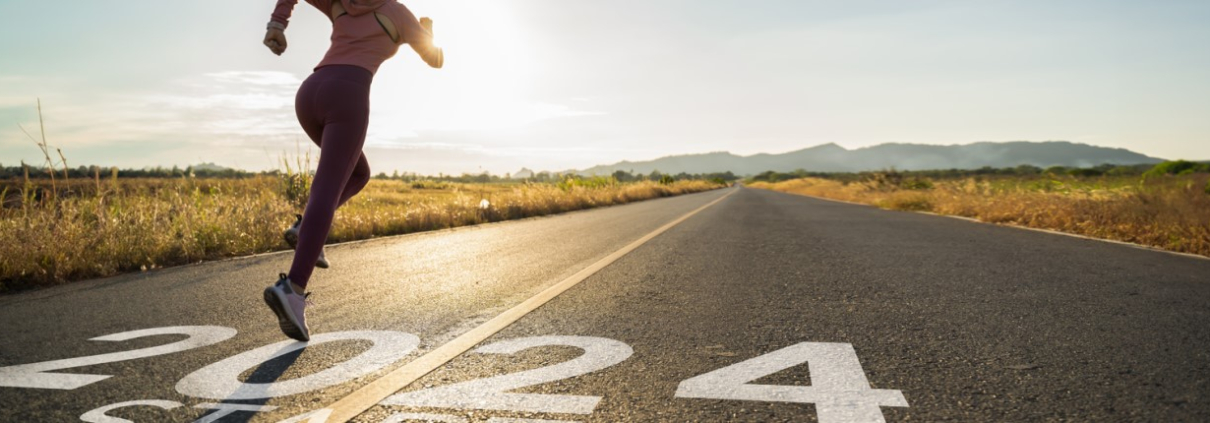 A person sprinting on a road with "2024" painted on the surface, indicating the year, with an early morning or late afternoon rural landscape in the background, featuring a clear sky and distant mountains.