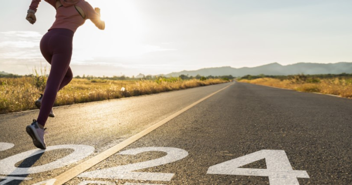 A person sprinting on a road with "2024" painted on the surface, indicating the year, with an early morning or late afternoon rural landscape in the background, featuring a clear sky and distant mountains.