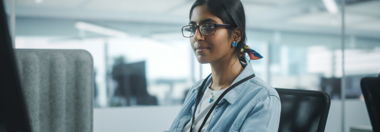 A focused young woman in glasses and a denim jacket working at a computer in an office setting.