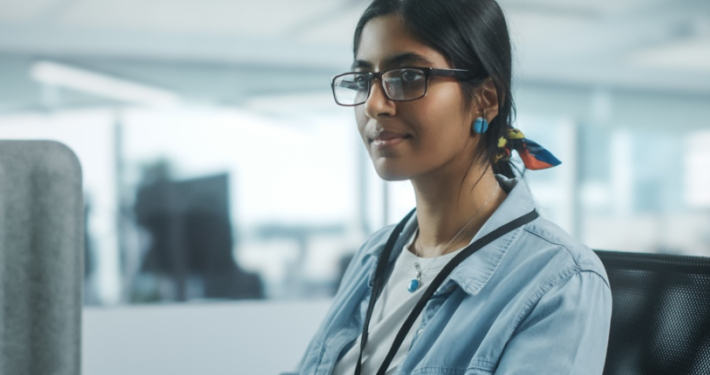 A focused young woman in glasses and a denim jacket working at a computer in an office setting.