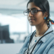 A focused young woman in glasses and a denim jacket working at a computer in an office setting.