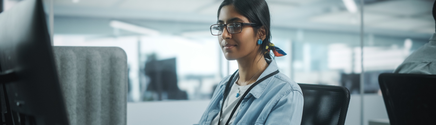 A focused young woman in glasses and a denim jacket working at a computer in an office setting.