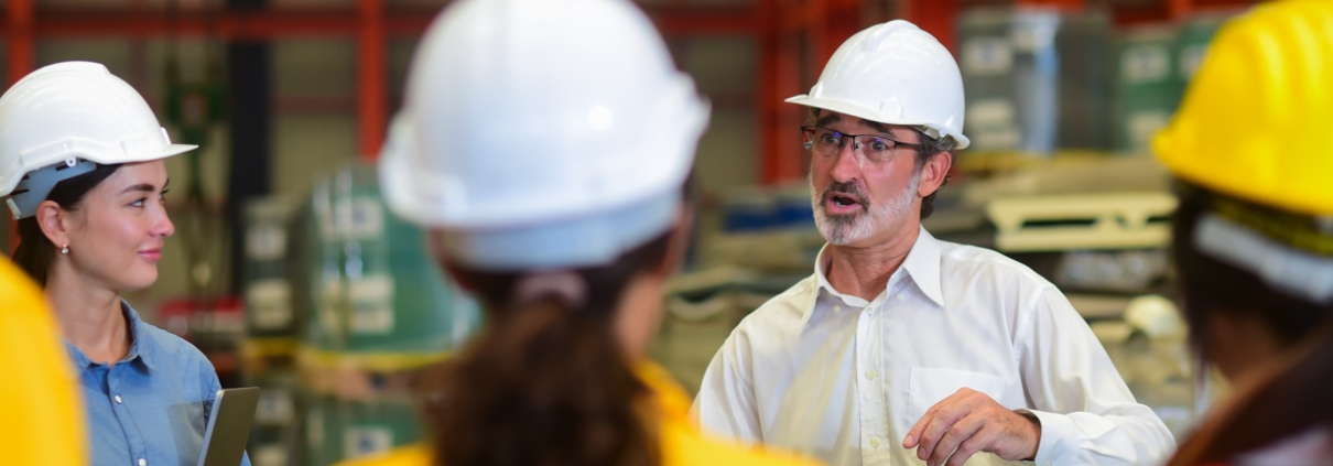 A group of workers wearing hard hats have a meeting in a warehouse