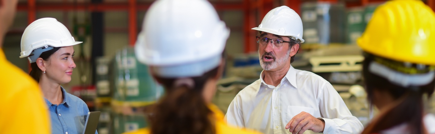 A group of workers wearing hard hats have a meeting in a warehouse