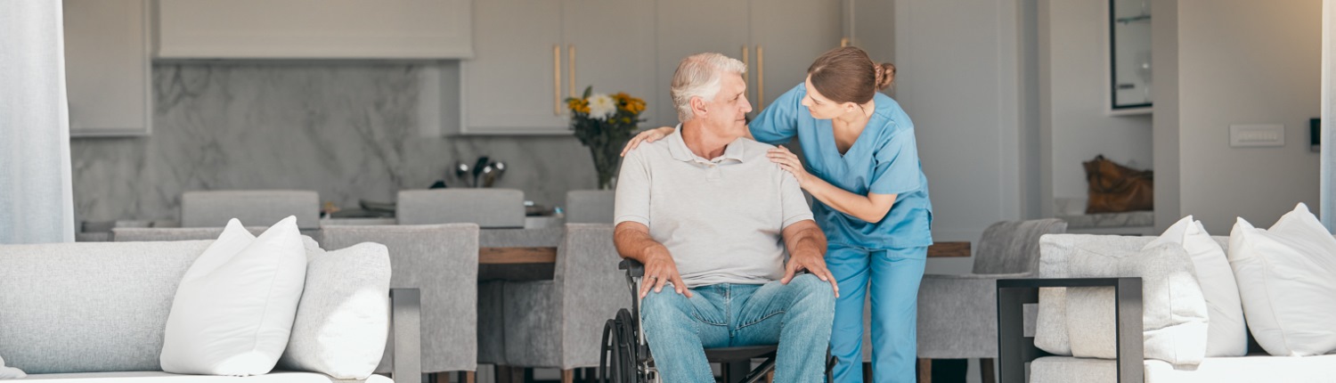 A nurse reassures a man in a wheelchair in a neutrally-decorated home setting