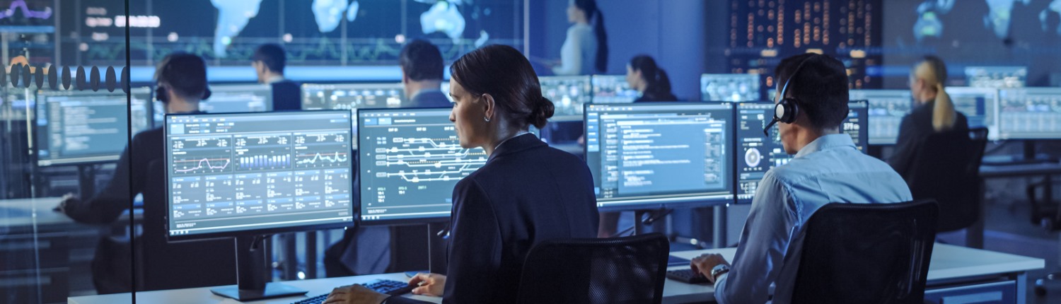 Workers sit in front of computer monitors displaying multiple data feeds, implying high-level monitoring or analytics work