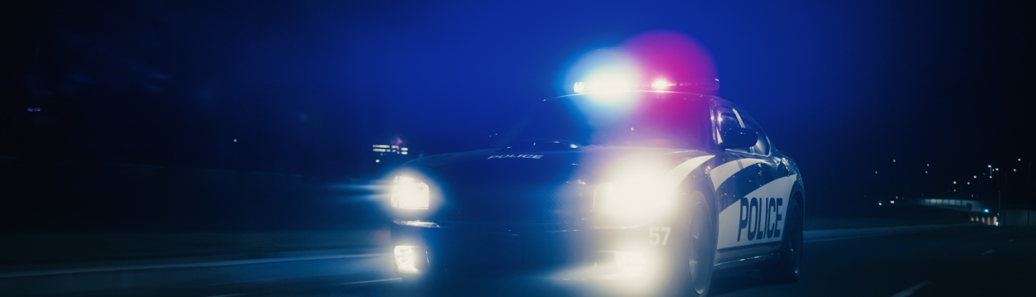 A police car drives down a street at night with its roof lights illuminated